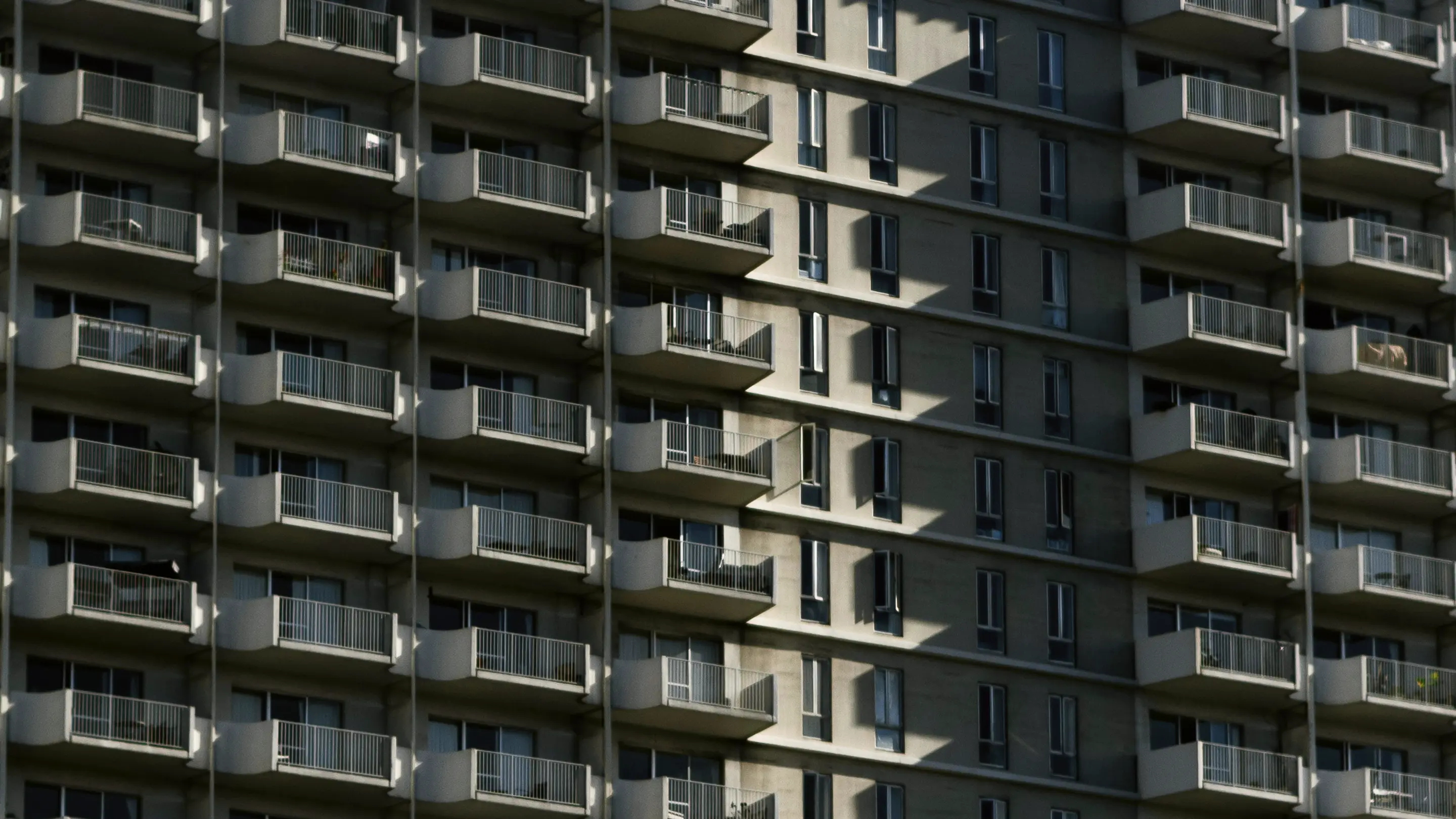 Фасад багатоповерхового житлового будинку з повторювальним малюнком балконів, великих вікон, бетонних колон та металевих перил у м'яких тонах бежу та сірого.