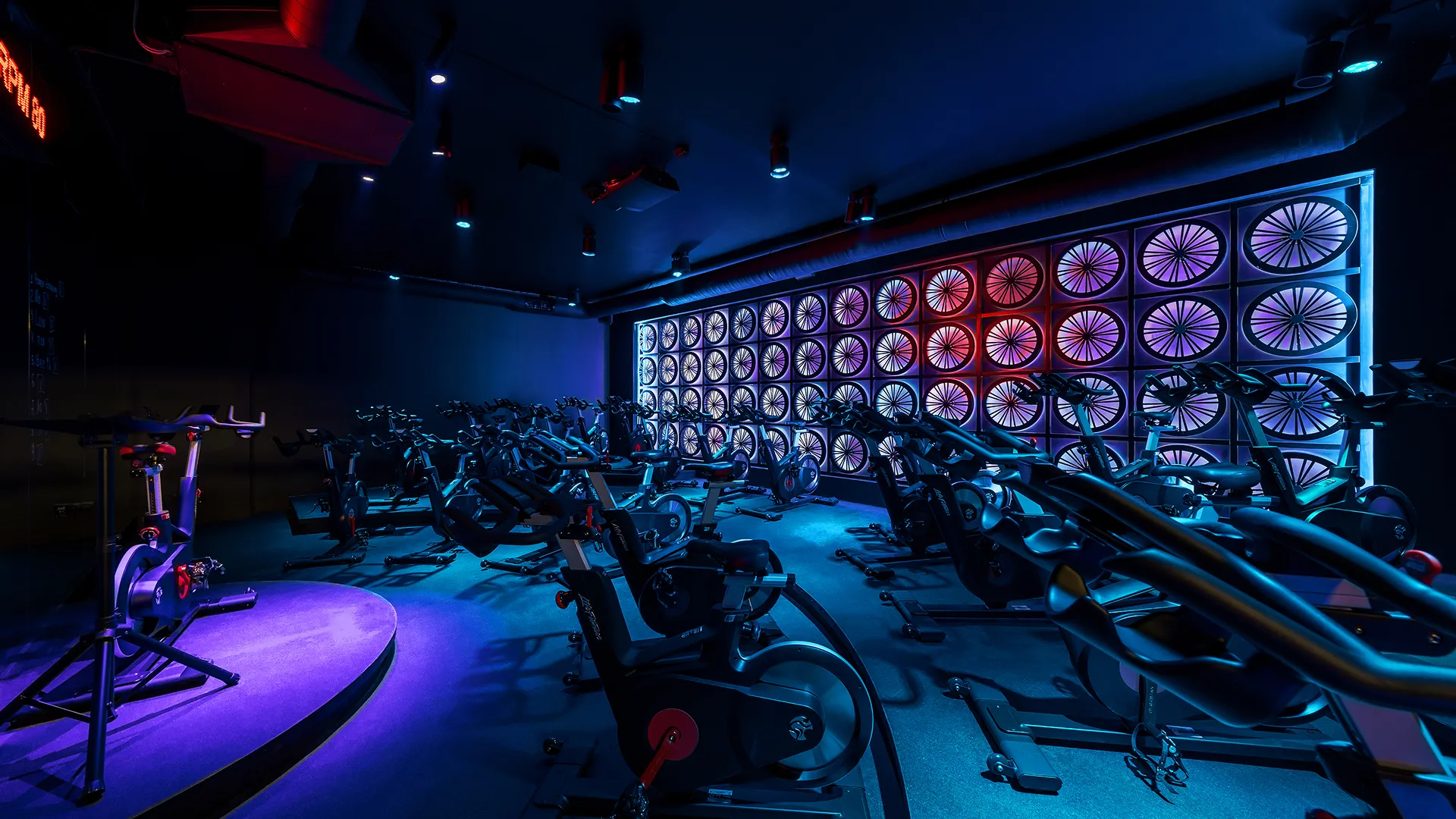 Внутрішній простір спортивного залу для велоспорту з неоновою підсвіткою: синім, фіолетовим та червоним світлом, рядами тренажерів для спінінгу, панеллю з світлових велосипедних коліс на стіні та мінімалістичним дизайном.