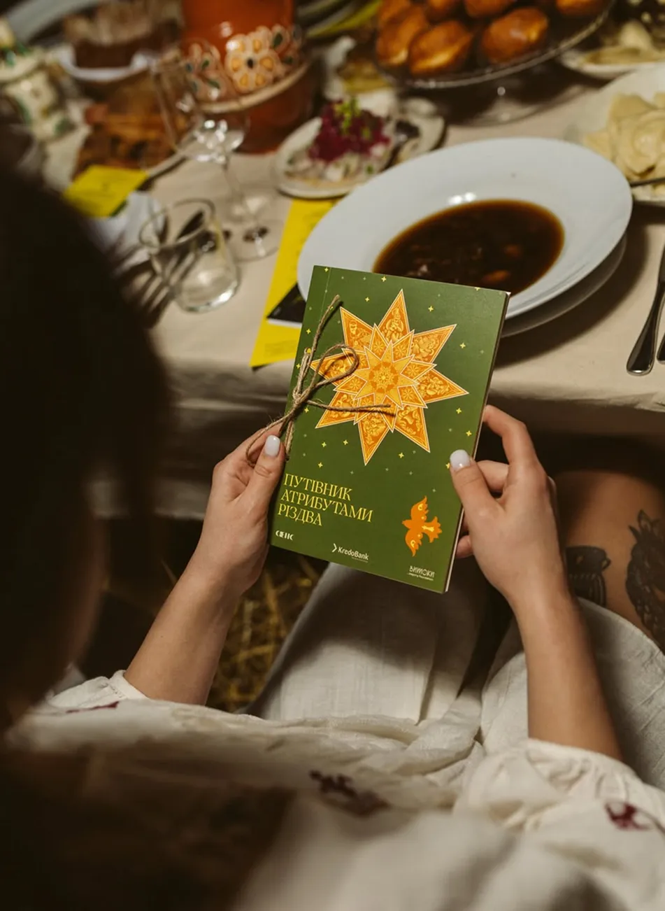 Людина тримає зелений буклет із жовтою різдвяною зіркою за столом із святковими стравами.