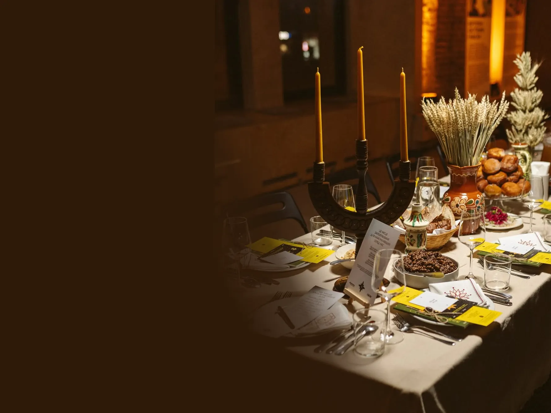 Святково накритий стіл в теплому освітленні: свічки у свічнику-тризубі, сніп пшениці в розписному керамічному глечику, пампушки, кутя. На місцях — меню, келихи для вина, лляна скатертина.