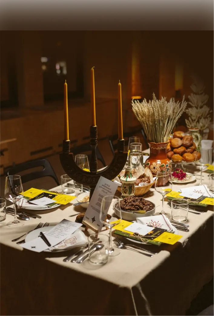 Святково накритий стіл в теплому освітленні: свічки у свічнику-тризубі, сніп пшениці в розписному керамічному глечику, пампушки, кутя. На місцях — меню, келихи для вина, лляна скатертина.